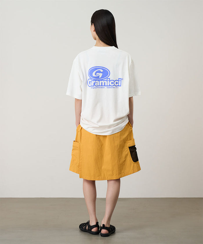 Rear view of a model wearing a white Gramicci '82 Tee with a blue graphic and yellow skort on a light tone background.