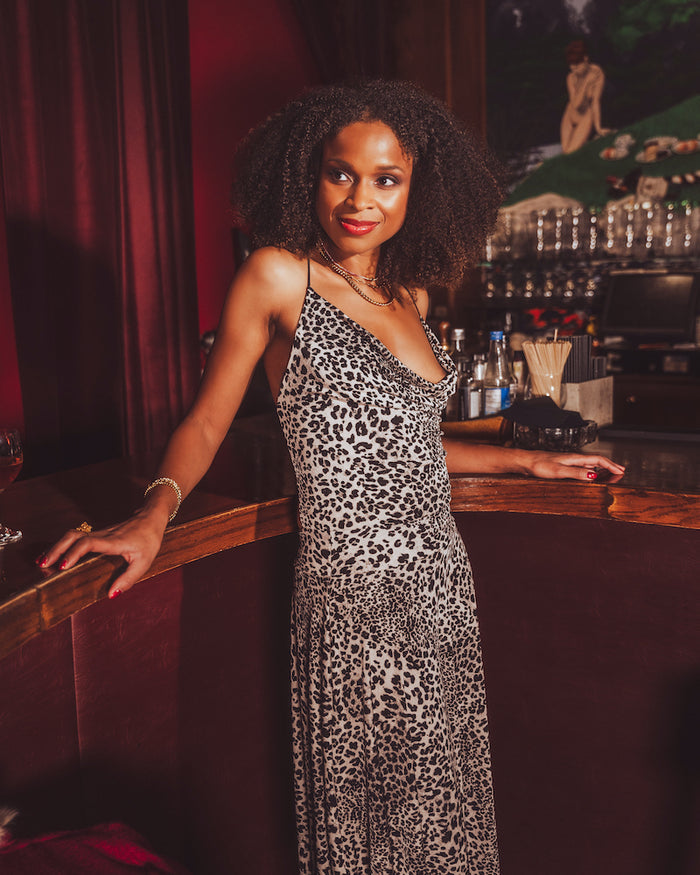 Woman in a  black and white leopard print dress standing at a 1970s style bar counter with a dark, possibly vintage interior.