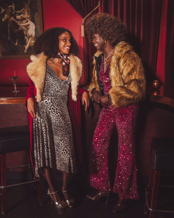 Two women in 1970s style glamorous outfits with fur coats standing in a  vintage room with red walls and a painting.