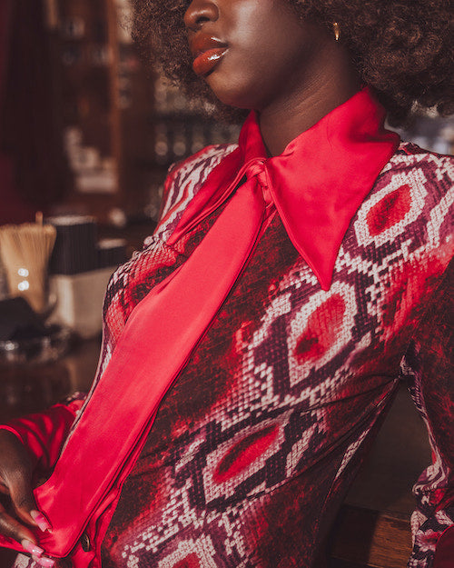 Person wearing a patterned outfit with a red satin bow tie in a blurred indoor setting
