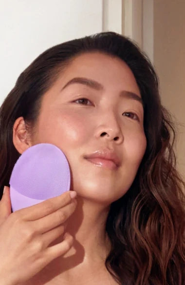 Woman using a lavender-colored LUNA™ 4 | FOREO facial cleansing device on her cheek for skincare and wellness.