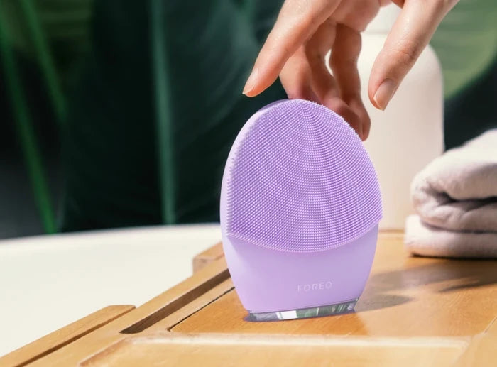 Hand holding a lavender LUNA™ 4 | FOREO silicone facial cleansing brush on a wooden surface with a folded towel and greenery in the background, showcasing a skincare wellness device.
