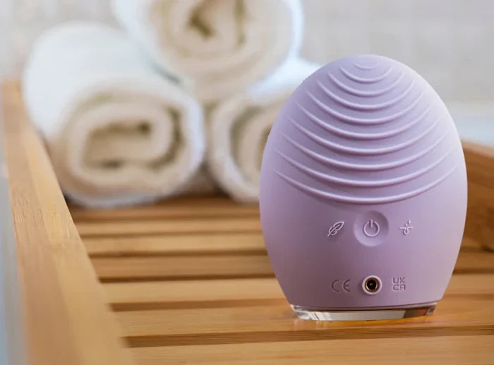 LUNA™ 4 | FOREO in soft lavender color placed on a wooden surface with rolled towels in the background, showcasing a sleek skincare and wellness device for facial massage.