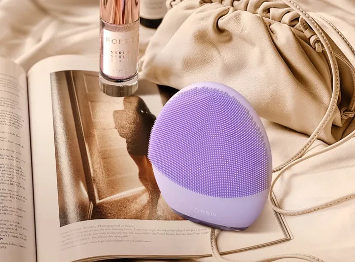 LUNA™ 4 | FOREO lavender silicone facial cleansing device placed on an open magazine with a beige handbag and skincare serum bottle in the background, suggesting a relaxing skincare routine.
