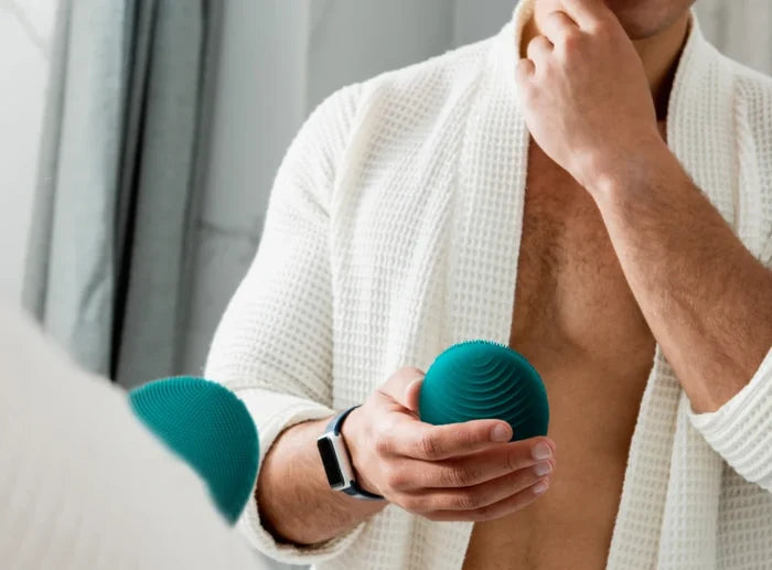 Man in a white robe holding a teal cleansing device in front of a mirror, demonstrating the skincare tool LUNA™ 4 MEN | FOREO for facial care and grooming.