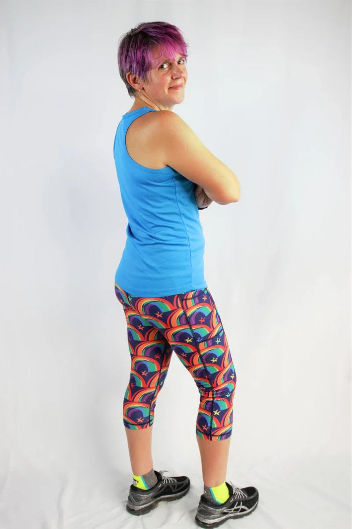 Woman with purple hair wearing a blue tank top and colorful rainbow print capris against white background