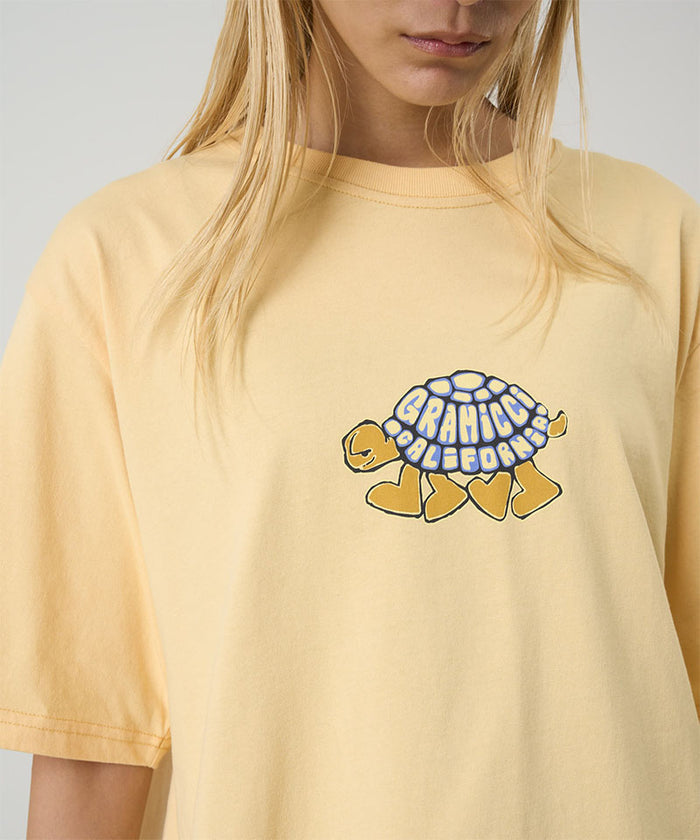 Model wearing a Gramicci mango Turtle Tee with a turtle graphic against a light tone background.